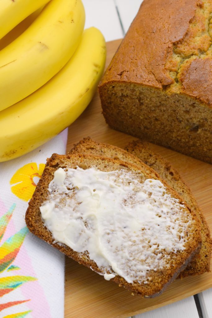 slices of banana bread with butter next to loaf of banana bread on wood board and bunch of bananas