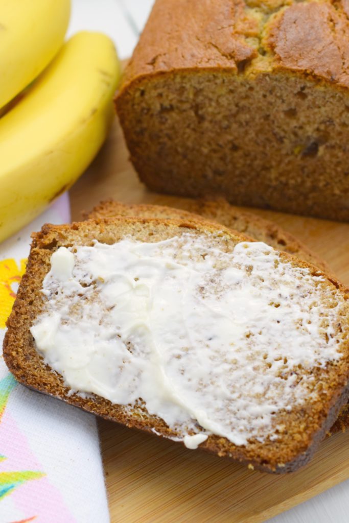 slice of buttered banana bread next to bunch of bananas and loaf on wood board